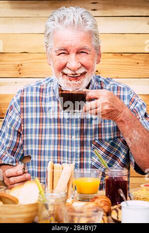 Allegro e divertente attivo uomo anziano che beve cioccolata calda con barba e baffi sporchi - felice pensionati che si godono la colazione - parete in legno di stile sullo sfondo - tavolo pieno di cibo Foto Stock