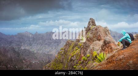 Uomo escursionista con zaino seduto sulla cima della montagna e guardando verso la valle. Splendido paesaggio arido sull'isola di Santo Antao, Cabo Verde. Foto Stock