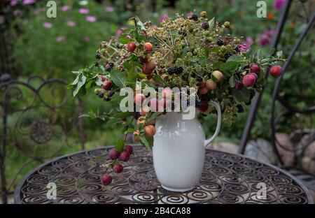 Primo piano d'autunno ancora vita con un bouquet di ramoscelli, bacche e piccole mele in una brocca bianca rustica, disposti su un tavolo vintage Foto Stock