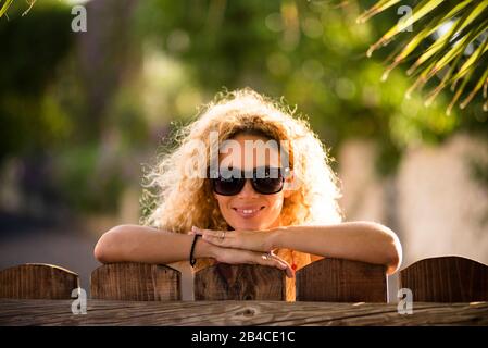 Ritratto di bella caucasica giovane bionda curly donna allegro guardando la fotocamera con luci del sole in background e defocused verde naturale sfondo esterno - felice età media signora concetto Foto Stock