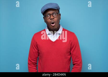 Bell'uomo africano americano nero in maglione rosso e cappello blu che guarda sorpreso. Studio girato su parete blu Foto Stock