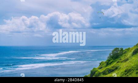 Parapendio volare fuori dalle colline erbose sopra Gunung Payung Beach nel nord-est di Bali una delle aree popolari per il parapendio. Foto Stock