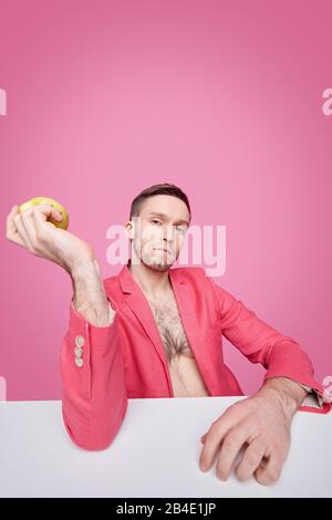 Bel giovane uomo in giacca rossa che tiene mela o qualche altra frutta mentre si rilassa alla scrivania davanti alla fotocamera su sfondo rosa Foto Stock