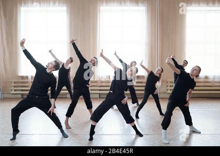 Gruppo di giovani ballerini attivi in magliette nere e pantaloni che allungano un braccio davanti a se stessi durante l'esercizio di danza Foto Stock