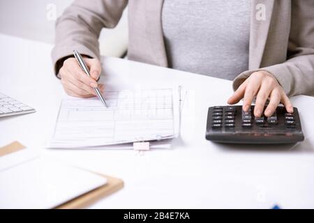 Mani di giovane ed elegante ragioniere con penna sopra documento finanziario premendo i pulsanti della calcolatrice mentre lavora da scrivania Foto Stock