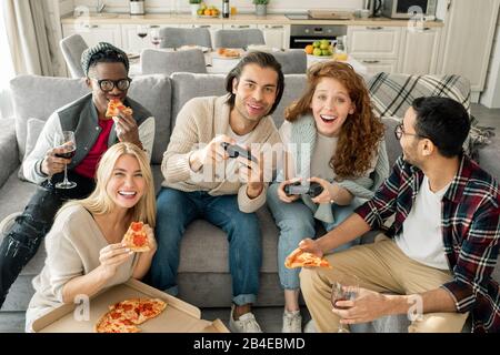 Gruppo di giovani amici interculturali che si godono la pizza, mentre due di loro che giocano a videogame in casa festa Foto Stock