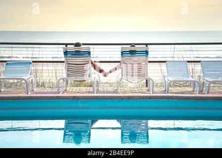 USA, Folrida, Daytona Beach, coppia in sedie da spiaggia accanto alla piscina, con le mani in mano, con vista sul mare