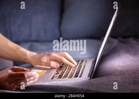 L'uomo ha le mani che tengono la carta di credito, pagano in linea e che immette il codice di sicurezza, usando il computer portatile e sdraiato sul divano di notte, fuoco morbido, spazio di copia Foto Stock