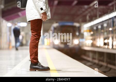 Primo piano di uomo in attesa treno alla stazione della metropolitana di mattina presto e va al lavoro, vestito di parka bianco, jeans rossi, stivali in pelle marrone, metropolitana pl vuoto Foto Stock