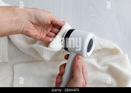 Mano dell'uomo che tiene un smacchiatore di pelucchi da un maglione bianco di lana o acrilico. Dispositivo elettrico dopo la pulizia e raccolta lanugine/lanugine, backgrou in legno chiaro Foto Stock