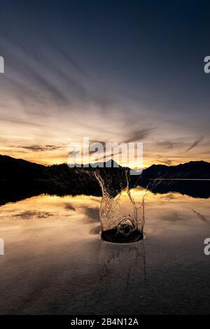 Una fontana d'acqua attraverso una pietra gettata nelle acque tranquille e limpide del Walchensee nelle Prealpi Bavaresi. Le montagne sullo sfondo. Foto Stock