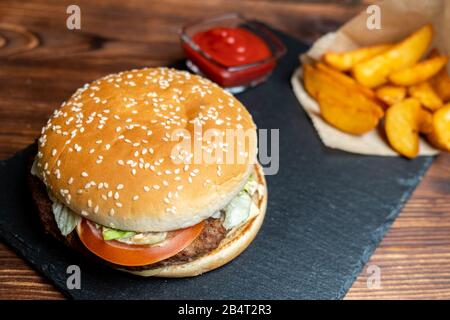 Burger in carta con patate in un rustico e ketchup e su ardesia, bordo nero e legno bruciato sfondo. Primo piano. Foto Stock