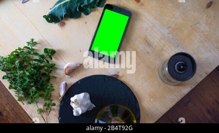 Momenti di vita quotidiana nella cucina mediterranea: Vista dall'alto a pianta piatta di uno smartphone con schermo verde vuoto su un tagliere di legno chiaro con bro Foto Stock