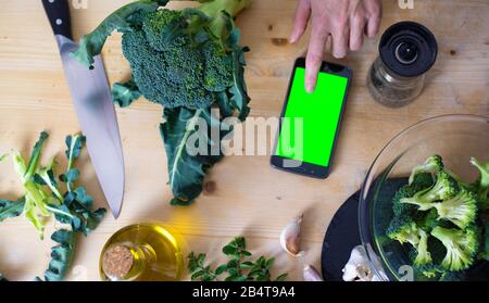 Momenti della vita quotidiana nella cucina mediterranea: Piatto vista della parte superiore della disposizione di una giovane cuoco femminile consultare il suo smartphone con schermo verde vuoto per trovare un Foto Stock