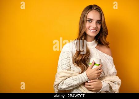 Una bella ragazza con pelle pulita e ricci in un maglione bianco si alza sorridendo con una tazza termica in mani su uno sfondo giallo Foto Stock