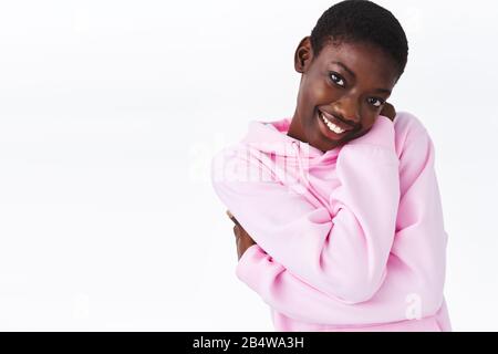 Concetto di cozyiness, tenerezza e bellezza. Adorabile giovane e romantica donna afro-americana abbracciare il proprio corpo, sorridendo amabile e guardando la fotocamera alluring Foto Stock