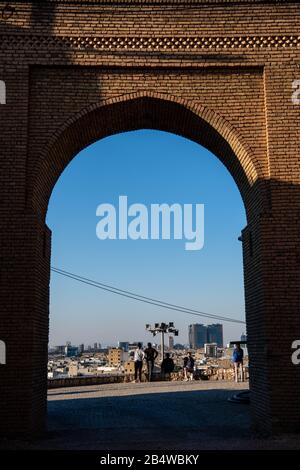 Iraq, Kurdistan Iracheno, Arbil, Erbil. Vista di Erbil al tramonto dall'interno della cittadella di Qalat. La vista è incorniciata dall'arco di ingresso della cittadella. Foto Stock