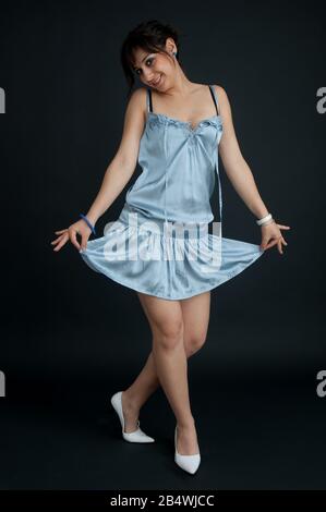 Bella ragazza bruna con i capelli raccolti, in abito azzurro che guarda la macchina fotografica sorridente e branchiante, su sfondo nero, scatto lungo Foto Stock