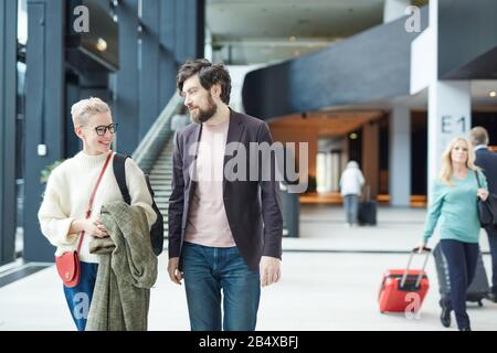 Giovane caucasico uomo e donna che chiacchierano mentre camminano lungo la sala dell'aeroporto, orizzontale medio lungo colpo Foto Stock