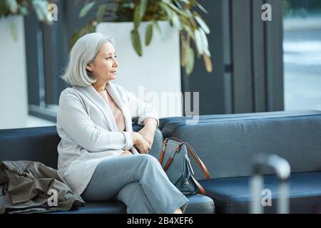 Ritratto orizzontale di donna anziana con i capelli grigi seduto da solo nella sala di partenza guardando lontano pensare a qualcosa, copia spazio Foto Stock