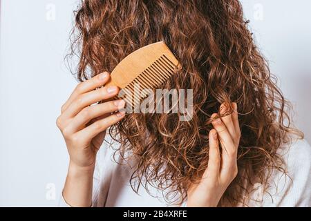 Primo piano giovane donna pettinare i capelli ricci con pettine in legno che si erge su sfondo bianco studio. Foto Stock