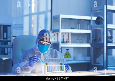 Colpo orizzontale di uno scienziato medico maschile irriconoscibile in indumenti protettivi seduti alla scrivania in un moderno laboratorio che tiene provetta con pericoloso l Foto Stock