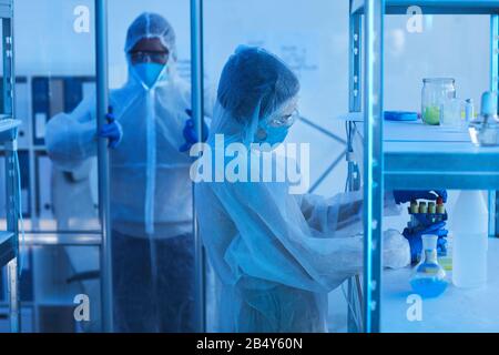 Medico maschile irriconoscibile in indumenti da lavoro protettivi apertura porta a sala da laboratorio, la sua collega femmina che prende provette da banco Foto Stock