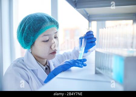 Ritratto di primo piano orizzontale di una giovane donna asiatica seria in camice da laboratorio che osserva la reazione chimica nella provetta Foto Stock