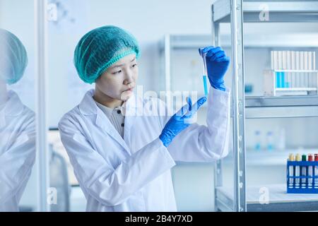 Colpo medio orizzontale di giovane lavoratore di laboratorio asiatico che indossa cappotto bianco, guanti di protezione e tappo che tiene la provetta con liquido blu Foto Stock