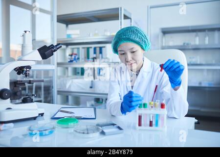 Ritratto orizzontale di giovane donna concentrata che indossa camice da laboratorio, guanti di protezione e cappello che esegue test medici, copia spazio Foto Stock