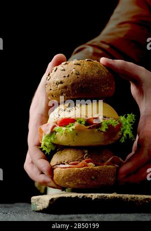 Le mani di una giovane donna stanno tenendo deliziosi hamburger croccanti con prosciutto affettato su un tagliere di legno vintage. Cibo scuro moody ancora vita. Foto Stock