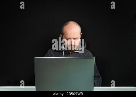 Uomo caucasico con cappuccio seduto dietro il computer portatile e digitando, sfondo nero Foto Stock