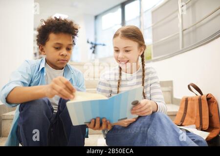 Serious Black boy e felice caucasica ragazza seduta insieme sulle scale leggere libro insieme, orizzontale medio ritratto shot Foto Stock