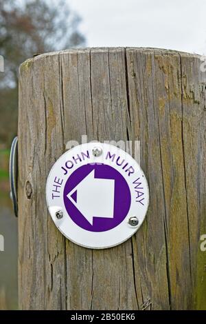 Cartello per la John Muir Way, un sentiero escursionistico a lunga distanza in Scozia. Su palo di legno con sfondo sfocato Foto Stock