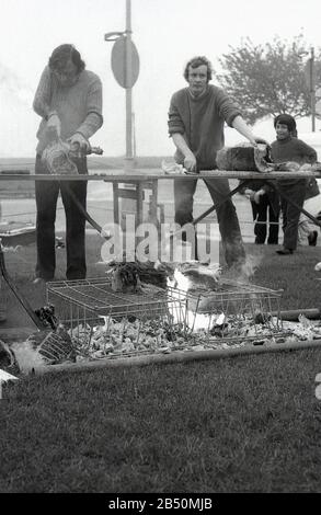 Anni '1970, storico, grandi pezzi di carne sono stati tagliati su un tavolo tressel e cucinati su un barbecue fai da te, una griglia metallica sopra un fuoco aperto sedeva sull'erba in un evento all'aperto, Inghilterra, Regno Unito. Foto Stock