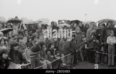 Anni '1970, storico, ombrelloni, come spettatori giovani e vecchi, molti indossano anaorak, si bagnano in piedi dietro una corda, guardando un concerto di musica all'aperto, Inghilterra, Regno Unito. Foto Stock