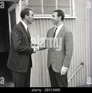 1960s, storico, allenatore di calcio del Charlton Athletic FC, Bob Stokoe che dà il benvenuto a un nuovo giocatore nel club, Charlton, South London, Inghilterra, Regno Unito. In piedi fuori dall'entrata, un capannone di metallo ondulato, entrambi gli uomini indossano giacche sportive a quadri e magliette e cravatte. Bob Stokoe fu il manager del club dal 1965 al 1967. Stokoe era famoso per la sua vittoria in fa Cup con il Sunderland, oltre ad essere un membro della squadra Newcastle FC che ha vinto la famosa coppa negli anni '1950 Foto Stock