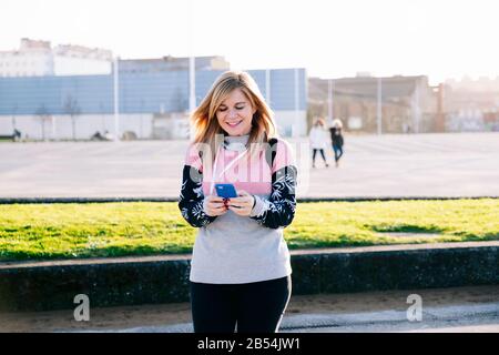 Una bella bionda donna caucasica usa un telefono cellulare con le mani che camminano lungo una passeggiata in città in una giornata di sole Foto Stock