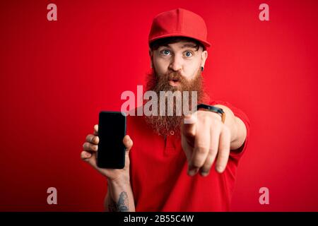 Red head Irish delivery man con smartphone Beard holding che mostra l'app sullo schermo e punta con il dito sulla fotocamera e su di te, il segno della mano, positivo e. Foto Stock