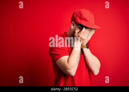 Giovane e bell'uomo che indossa occhiali e tappo rosso su sfondo isolato con triste espressione che copre il viso con le mani mentre piange. Depressione Foto Stock
