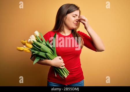 Giovane donna bionda che tiene un bouquet romantico di tulipani fiori su sfondo giallo che odorano qualcosa di puzzolente e disgustoso, odore intollerabile, tenere Foto Stock