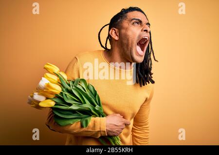 Giovane afroamericano afro romantico uomo con temute serrature che tengono bouquet di tulipani gialli arrabbiati e pazzi uroso urlando frustrato e furioso, gridando spirito Foto Stock