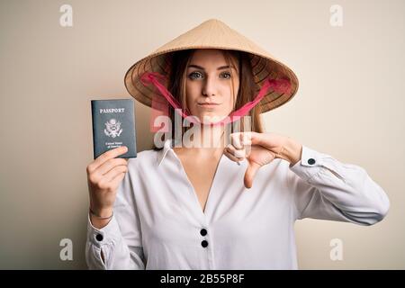 Giovane bella donna turistica redhead indossando cappello tradizionale asiatico tenendo passaporto usa con faccia arrabbiata, segno negativo che mostra antipatia con i pollici Foto Stock