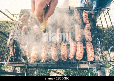 Giovane uomo che cucina carne sul barbecue - Chef mettere alcune salsicce di maiale alla griglia nel parco all'aperto - Concetto di mangiare barbecue all'aperto durante l'estate - Sof Foto Stock