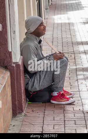 Mendicante maschile seduto sul marciapiede di fronte al negozio, con scarpe rosse e un cappuccio di lana, tenendo un cartello di cartone su state Street, Santa Barbara, CA Foto Stock