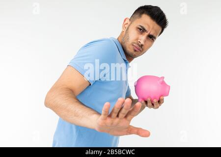 giovane uomo in una t-shirt blu tiene una banca piggy e disaccetta mostrando il gesto di rifiuto su uno sfondo bianco studio Foto Stock