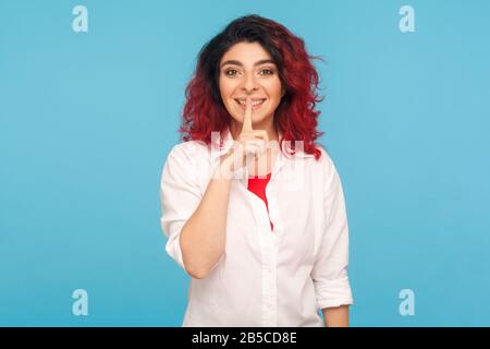 Shh, mantenere la calma! Ritratto di donna hippster positiva con capelli rossi fantasia in camicia bianca tenendo il dito sulle labbra, arrossendo e mostrando il gesto di silenzio. In Foto Stock