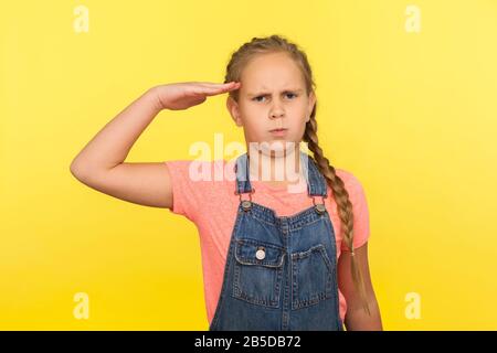 Sì, Signore! Ritratto di responsabile obbediente bambina con treccia in denim tuta saluting e guardando la fotocamera con rispetto, bambino patriottico. Indoo Foto Stock