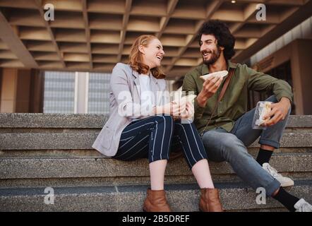 Sorridente giovane uomo e bella donna mangiare sandwich mentre in una conversazione seduto sulle scale Foto Stock