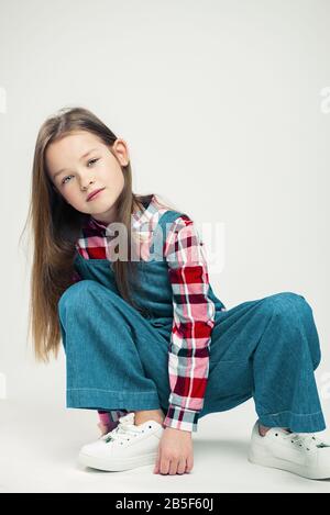 simpatica bambina seduta sul pavimento. Indossate un abito in denim, una camicia in plaid e un paio di sneaker bianche. Studio fashion photography. Moda bambino Foto Stock